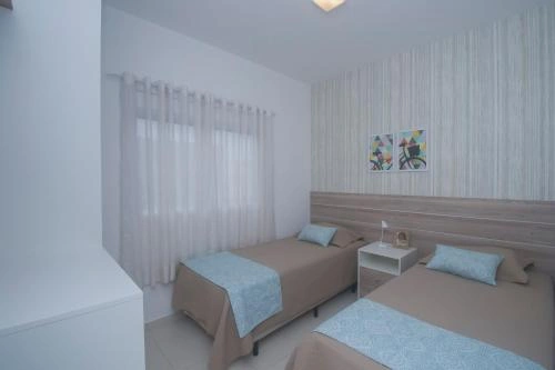 Twin bedroom with two single beds featuring tan covers and blue sheets, white curtains, a shared bedside table, and a white textured accent wall.