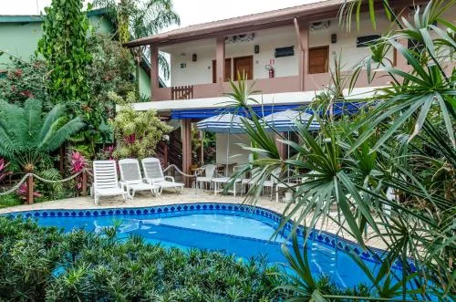 View of a tropical resort area featuring a blue tiled swimming pool, white sun loungers, lush green foliage, and a two-story building with a covered balcony.