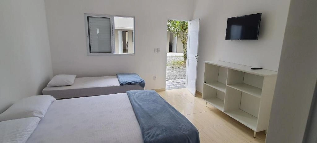 Spacious white guest bedroom with two twin beds, TV, white shelving unit, and a door opening to a sunny outdoor gravel area.