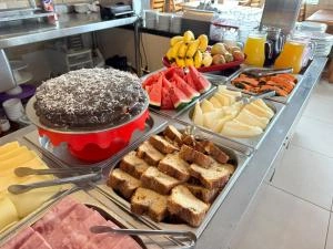 Abundant morning breakfast spread featuring a large chocolate coconut cake, fresh slices of watermelon, bananas, cheese, and bread rolls.
