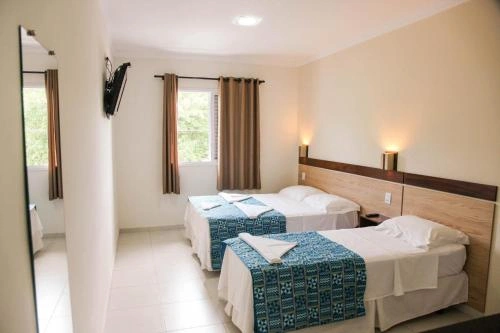 Interior view of a bright twin bedroom with two single beds, white bedding, patterned teal bed runners, brown curtains, wall-mounted TV, and warm bedside lamps.