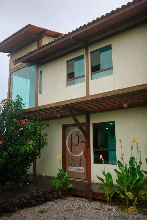 Low-angle view of a modern two-story building with a wood and glass facade, an outdoor wooden deck, and a painted sign on the door reading "Casa do Pedrinho Noronha" surrounded by plants.