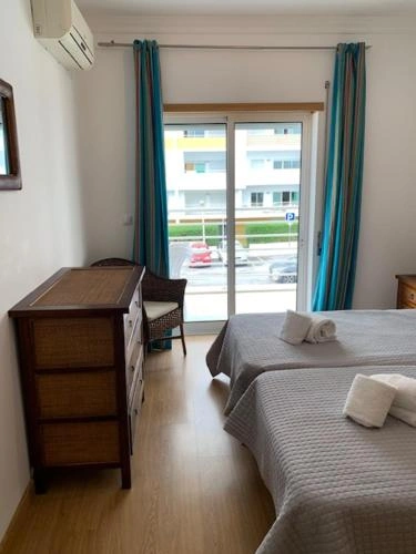 Twin bedroom with two grey beds topped with folded towels, a wooden dresser, teal curtains, and sliding glass doors showing a street view outside.