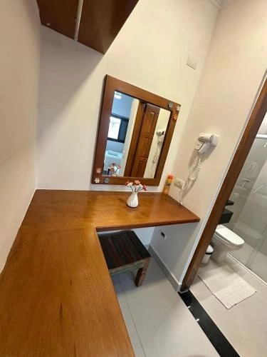 Interior view of a bathroom vanity featuring a wooden countertop, framed mirror, small stool, wall-mounted hairdryer, and a white vase with flowers.