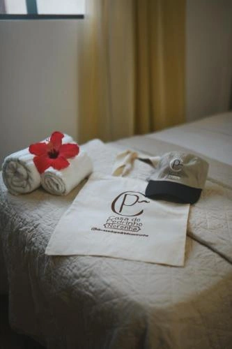 Close-up view of a bed with a textured beige bedspread, rolled white towels with a red flower, a branded canvas tote bag, and a small pouch resting on the bed.