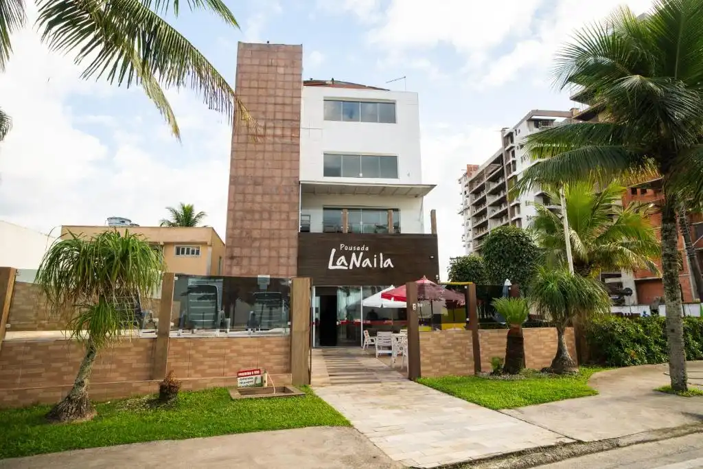 Full street-level view of Pousada La Naila showing the main glass entrance, mixed brick and white architecture, and surrounding palm trees.