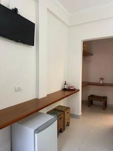 Interior corner of a hotel room featuring a wooden countertop, a white mini-fridge beneath it, a wall-mounted TV, and a glimpse into an adjacent vanity area.