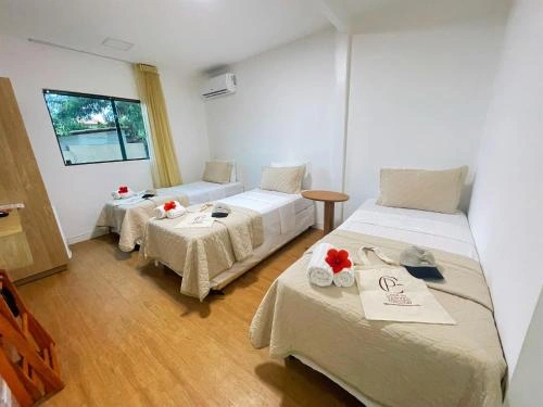 Wide-angle interior view of a bright bedroom with wooden floors, three single beds with matching tan bedding, white pillows, rolled towels with red flowers, and a window overlooking greenery.