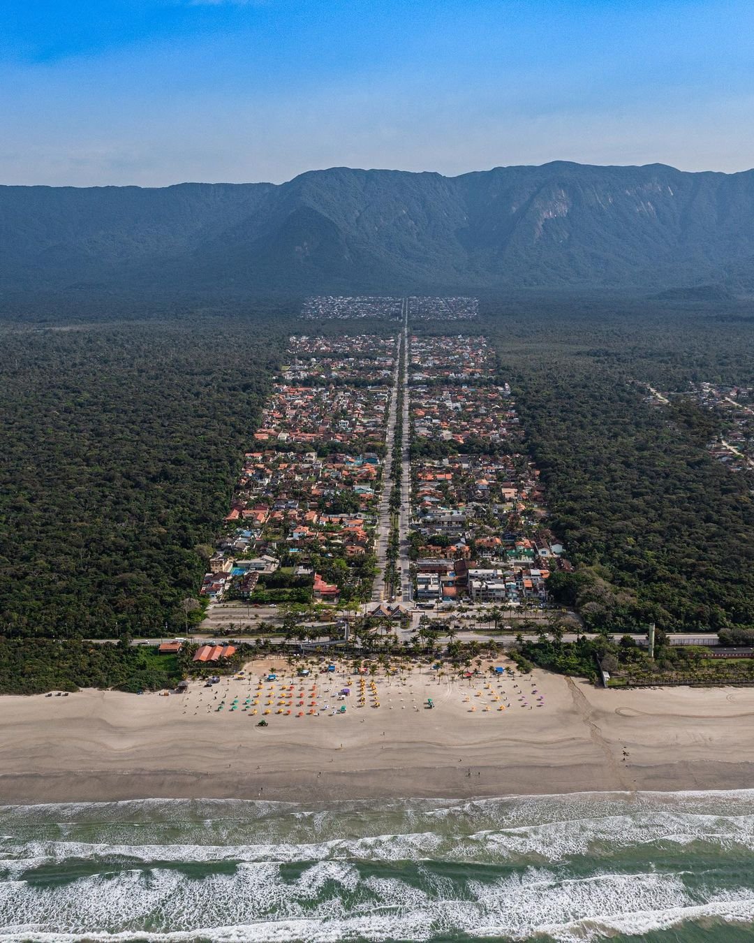 Aerial drone view of a narrow, rectangular coastal town bordered by dense forest and mountains, featuring a main central road, a sandy beach with colorful umbrellas, and breaking ocean waves in the foreground.