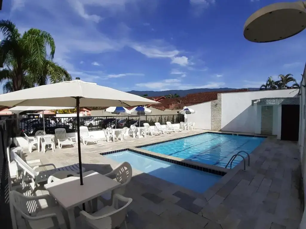 Spacious outdoor pool area with two rectangular swimming pools, white tables, umbrellas, and lounge chairs under a bright blue sky.