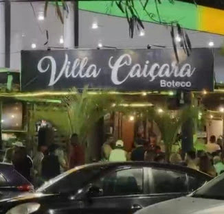Large illuminated "Villa Caicara Boteco" storefront sign with a busy crowd gathering outside at night.