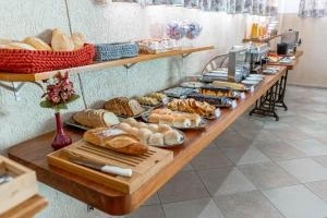 Wide view of a wooden hotel breakfast buffet counter featuring fresh bread rolls, sliced cakes, juices, and a coffee machine.