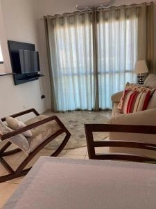 Relaxing indoor hotel lounge area featuring wooden rocking chairs, a beige sofa with red striped pillows, and a wall-mounted flat-screen TV.