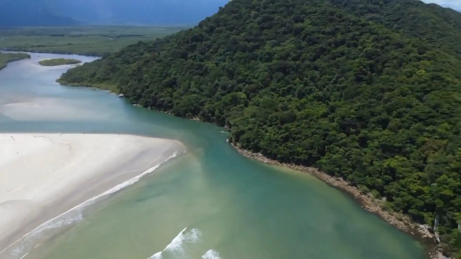 Aerial perspective of a green-tinted river entering the ocean, flanked by a wide white sandy beach on the left and a steep, forested mountain on the right.