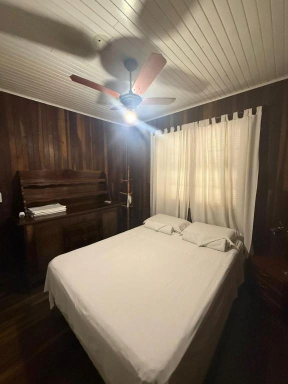 Cozy bedroom with dark wood-paneled walls, a white bed, a vintage wooden desk, white curtains, and a ceiling fan with a bright light fixture.