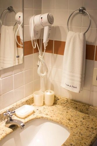 Bathroom counter displaying a white wall-mounted hairdryer, folded towel, two wrapped soap cups, and a granite sink with a chrome faucet.