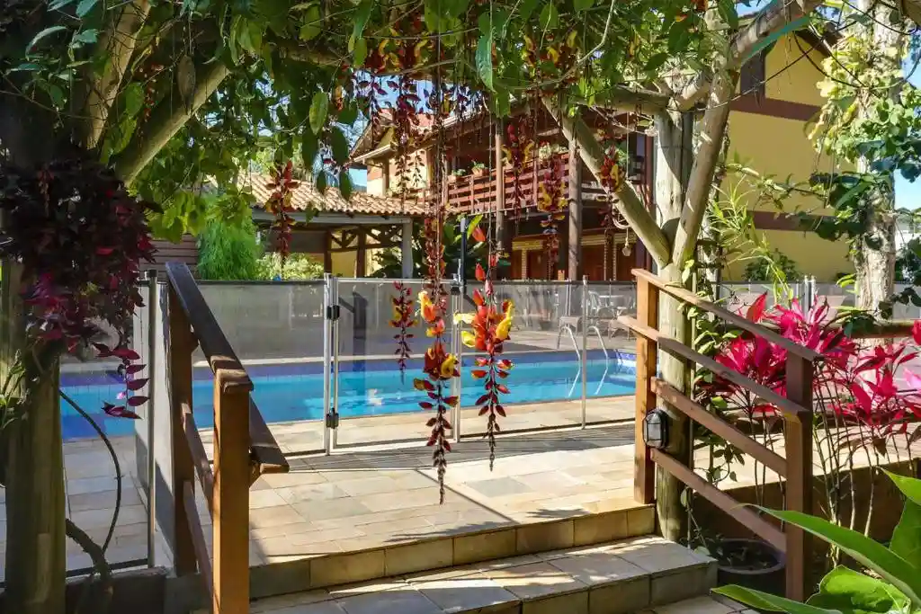 Garden path leading through wooden gates and lush, colorful plants to a bright blue swimming pool with a glass safety fence.