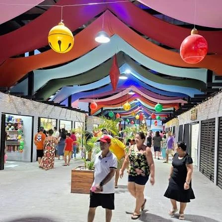 A colorful indoor market corridor decorated with vibrant, flowing ceiling streamers, large hanging ornaments, and shoppers walking along the tiled floor.