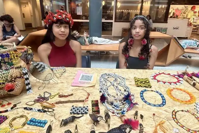 Two young women wearing traditional feathered headdresses and beaded jewelry sit behind a table displaying intricate woven baskets, beaded necklaces, and feather art.