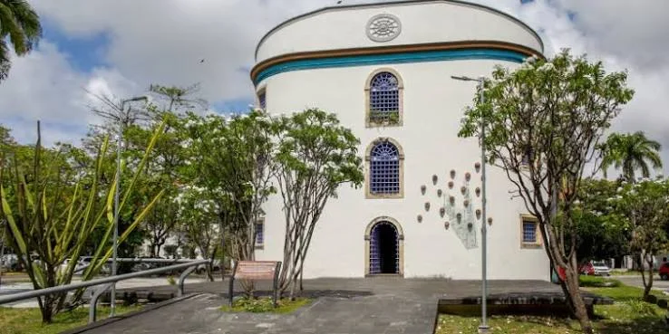 A striking, white cylindrical building featuring arched windows with blue stained glass, dark teal accents, and lush surrounding greenery.
