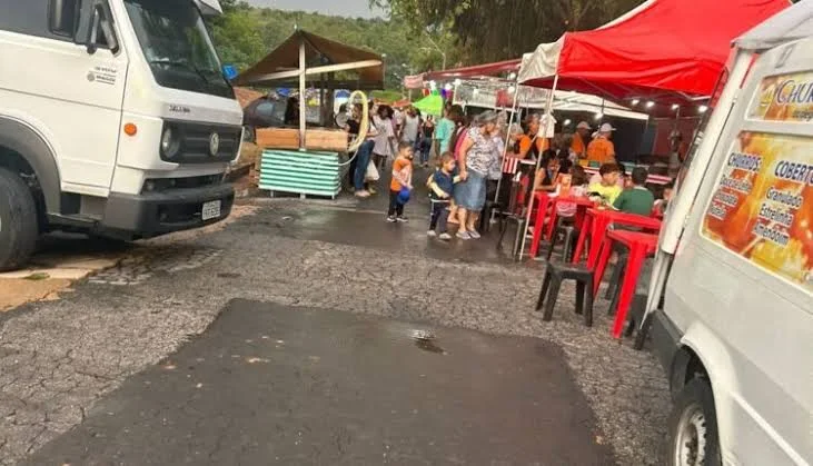 A lively outdoor food area featuring red canopies, red plastic chairs and tables, food trucks, and a crowd of people enjoying the market atmosphere.