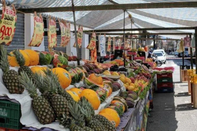 A sunny outdoor market stall with hand-written price signs, displaying a prominent arrangement of stacked pineapples, melons, and other fresh tropical fruits.