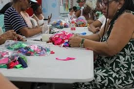 A group of people sitting together at a long white table, actively working on colorful, handmade crafts and beaded items.