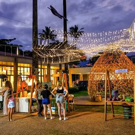 A magical evening outdoor market featuring overhead string lights, palm trees, a large woven wooden igloo structure, and a warm, inviting atmosphere.