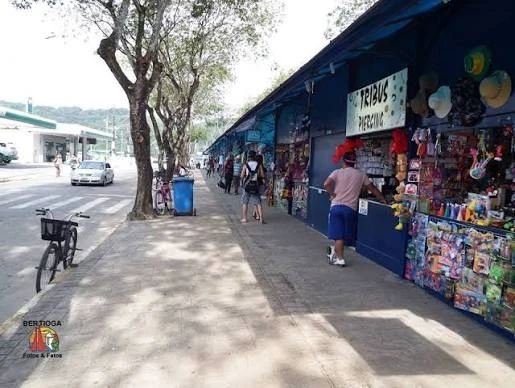 A row of blue outdoor shop stalls lining a street, displaying colorful hats, beachwear, and souvenirs, with a sign reading "Trabalho Artesanal" (Artisan Work).