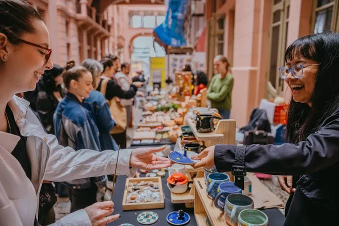 A smiling vendor handing a small blue ceramic plate to a happy customer at a vibrant outdoor flea market stall filled with pottery.