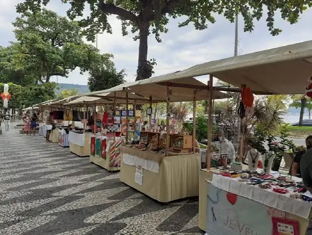 A long row of beige canopies set up on a wave-patterned mosaic sidewalk near the beach, selling jewelry and handmade goods.
