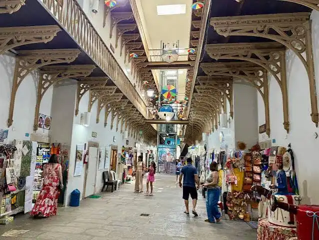 The interior of a grand, historic market hall featuring ornate iron archways, a high glass skylight, and shoppers browsing permanent vendor stalls.