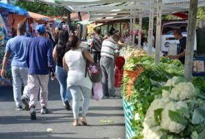 A bustling outdoor market aisle lined with fresh produce, showing shoppers walking past bright green cauliflower and leafy greens.