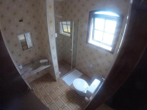 Private bathroom in a Pousada suite featuring a glass shower stall, toilet, sink with mirror, patterned wallpaper, and a frosted window for natural light.