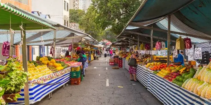 A vibrant open-air market lined with blue and white striped stalls under green awnings, packed with piles of fresh, colorful tropical fruits and shoppers.