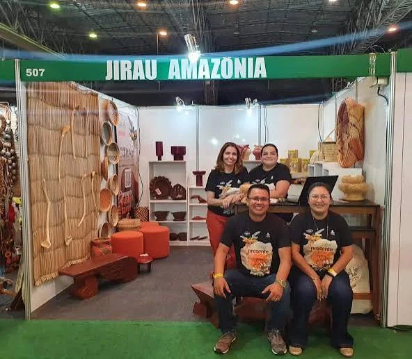 Four smiling people posing for a photo inside their "Jirau Amazônia" trade booth, which features wooden furniture, woven crafts, and pottery.
