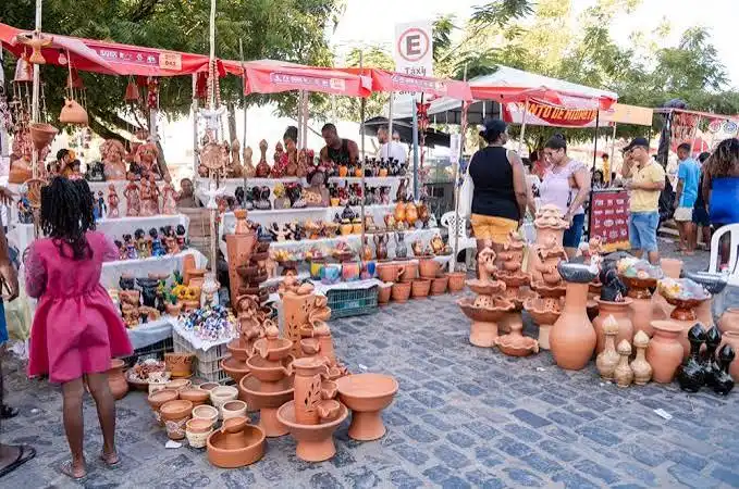 An outdoor market filled with red clay pottery, terracotta vases, and carved figurines, with shoppers browsing the displays under red awnings.