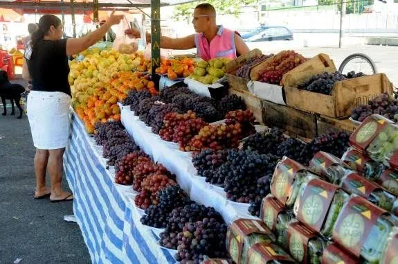 A market vendor handing a bag of fruit to a customer at a blue and white striped stall covered in neatly arranged bunches of grapes and yellow melons.