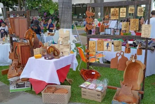 A market stall displaying beautiful handcrafted wooden furniture, cutting boards, spoons, and other carved wooden kitchen items on white-covered tables.