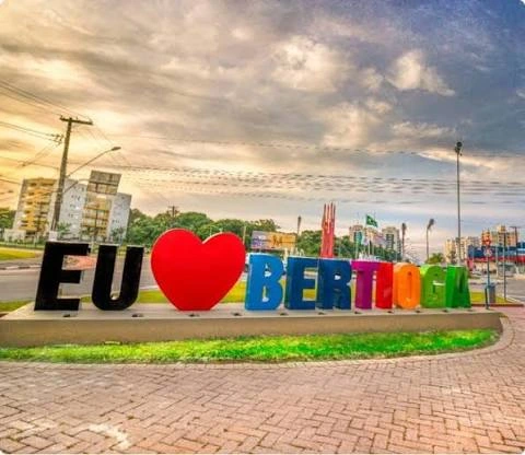 A colorful, oversized "EU ♥ BERTIOGA" sign made of giant, individual 3D letters placed on a landscaped grassy median against a dramatic, cloudy sky.