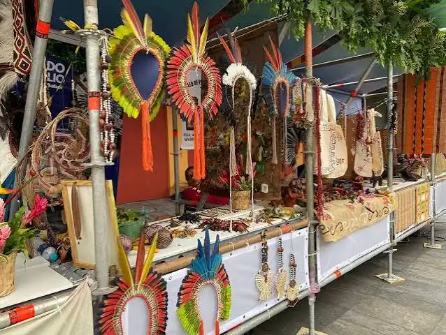 A vibrant market display of colorful, traditional indigenous feather headdresses and accessories hanging vertically from a metal rack.