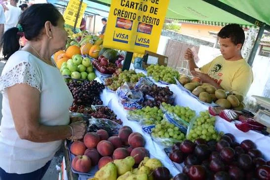 A vendor selling fresh fruits to a customer under a green canopy, with bright yellow signs displaying "Cartão de Crédito e Débito" (Credit and Debit Cards) and Visa logos.