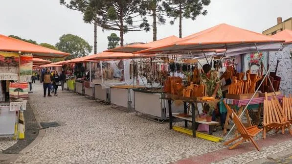A paved market walkway lined with a long row of bright orange canopy tents, selling wooden goods and handmade crafts under tall trees.