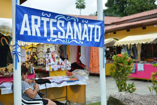 A bright blue "ARTESANATO" (handicrafts) banner hanging over a colorful outdoor stall displaying lacework, clothing, and handmade souvenirs.