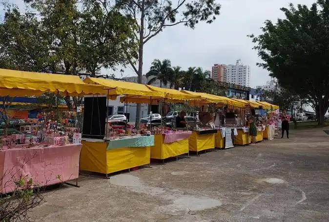 A row of temporary market stalls with bright yellow awnings and pink/white table covers, set up in a paved outdoor area surrounded by trees.