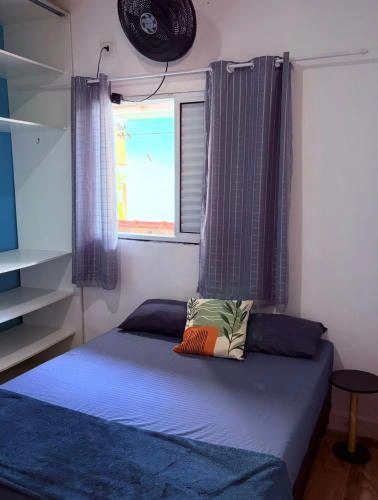 A simple bedroom featuring a double bed with blue bedding, a geometric accent pillow, gray curtains, and a wall-mounted fan.