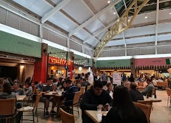 A busy mall food court bustling with diners beneath an arched glass ceiling, complete with a large Christmas tree and holiday decorations.