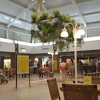 A relaxing indoor seating area within a shopping mall, shaded by a large suspended artificial tree canopy, with decorative street lamp fixtures and benches.