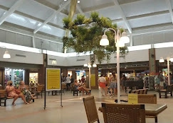 A spacious indoor mall seating area featuring wicker chairs, tables, and a central hanging garden structure providing shade.