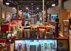 A retail display kiosk featuring an organized arrangement of L'Occitane skincare products, lotions, and gift sets in a brightly lit mall corridor.